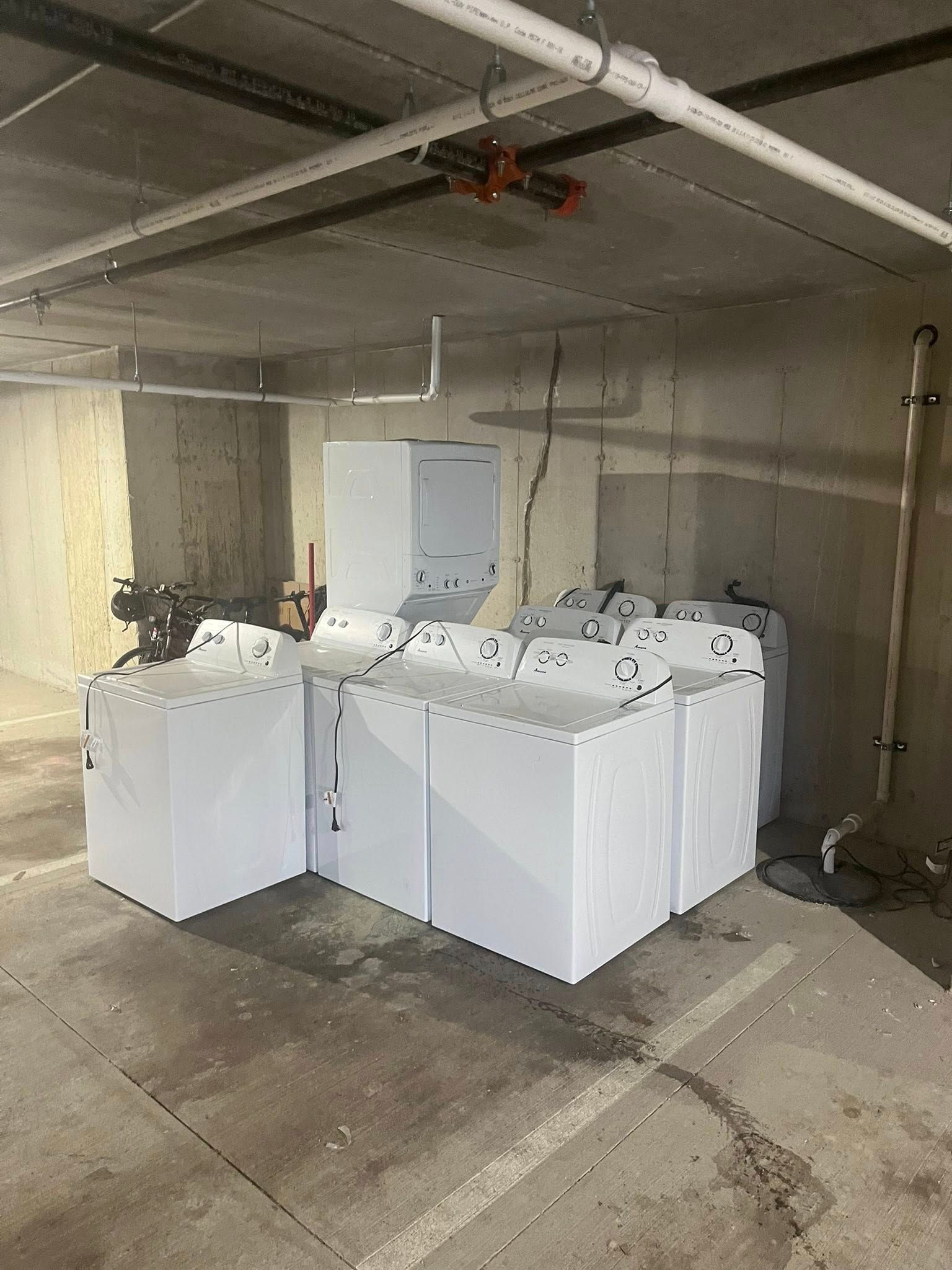 Cluster of white appliances or cabinets in a bare concrete basement with exposed ceiling pipes.