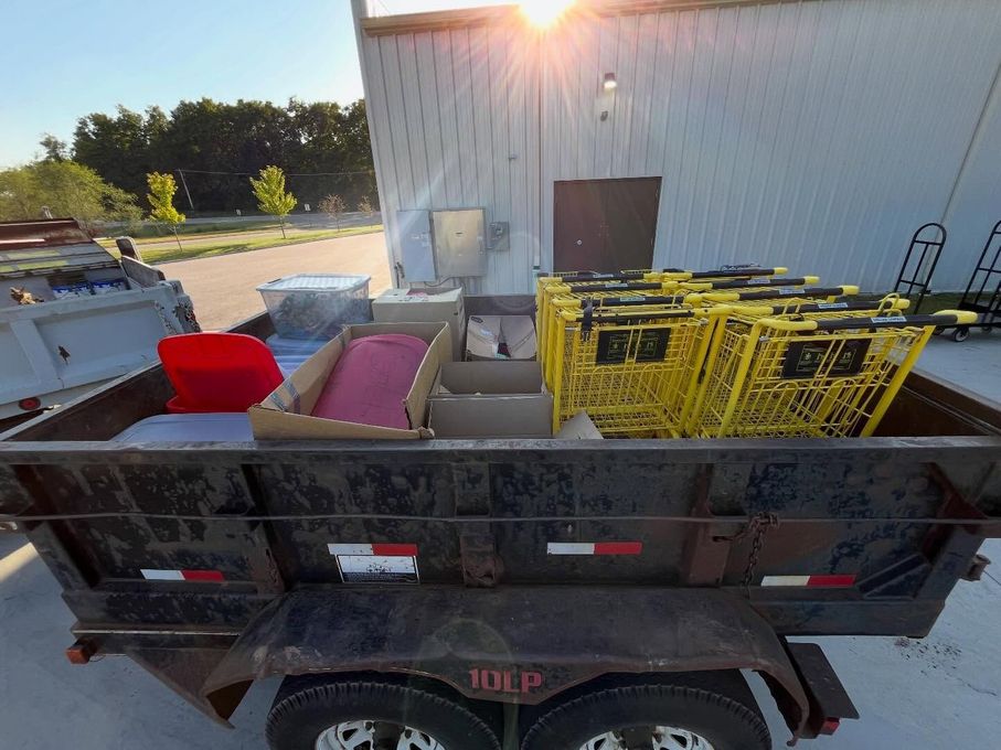 Trailer loaded with red chair, pink mattress, and yellow carts beside a white building at sunset
