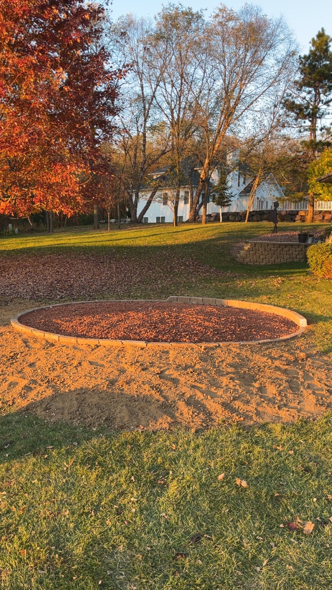 Autumn garden with a circular flower bed, fallen leaves, and a white house in the background.