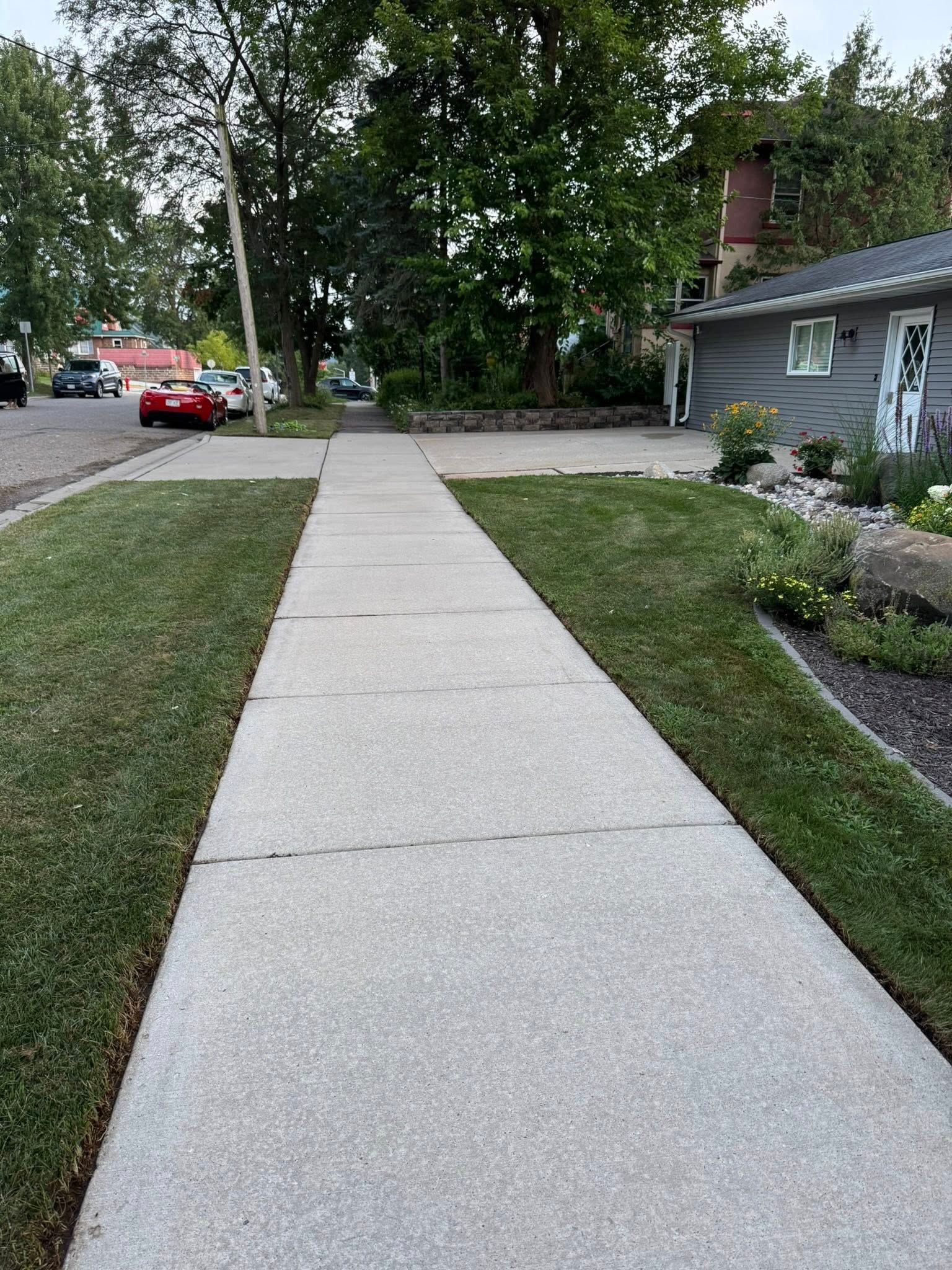 Sidewalk beside a house and lawn, with trees, parked cars, and a red car at the street corner.
