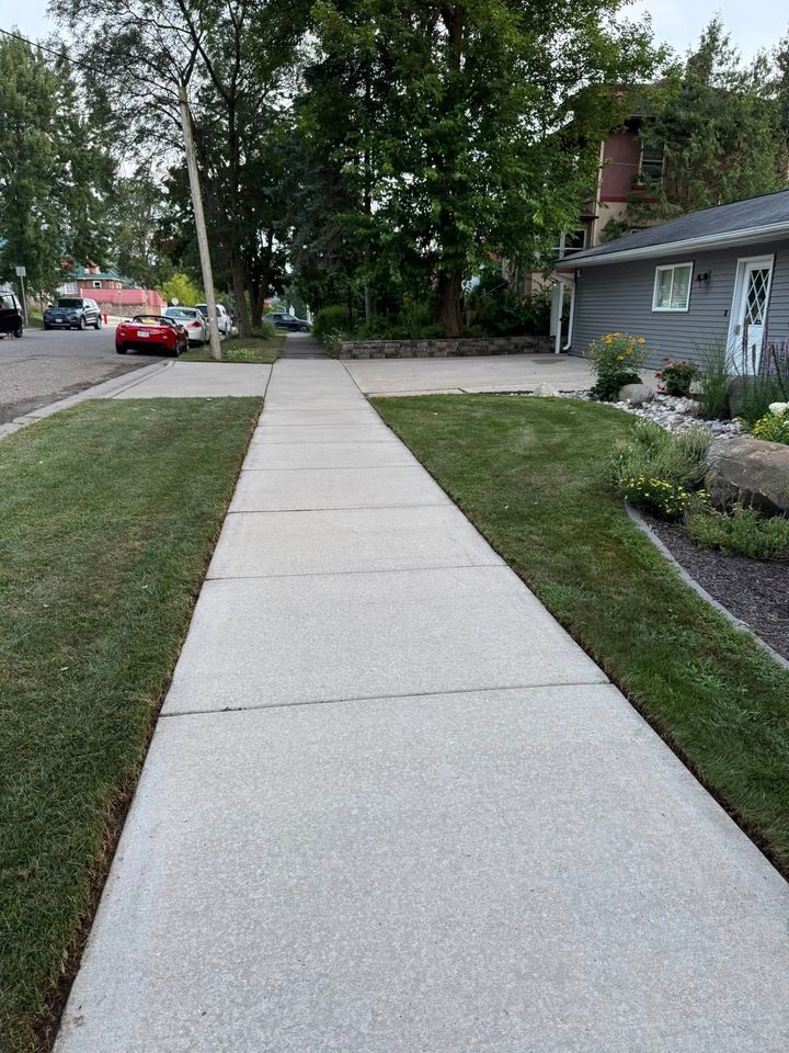 Sidewalk beside a house and lawn, with trees, parked cars, and a red car at the street corner.