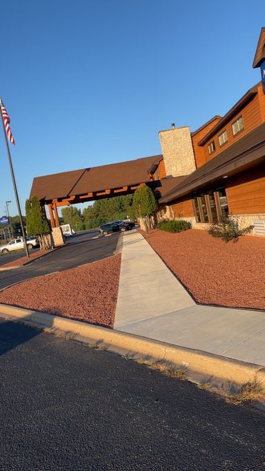 Lodge-style building entrance with a covered walkway, stone chimney, and landscaped sidewalk under a clear blue sky