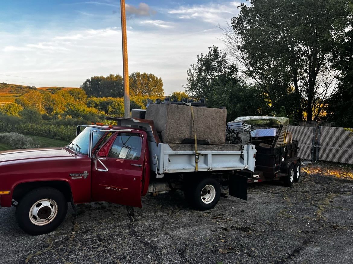 Red pickup truck loaded with debris on a gravel lot at dusk, with trees and a fence in the background