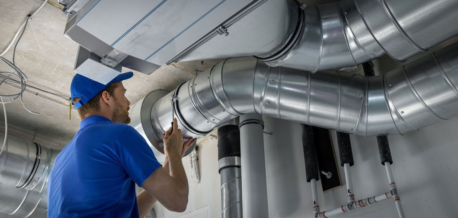 Man in blue uniform working on ductwork near a ceiling.