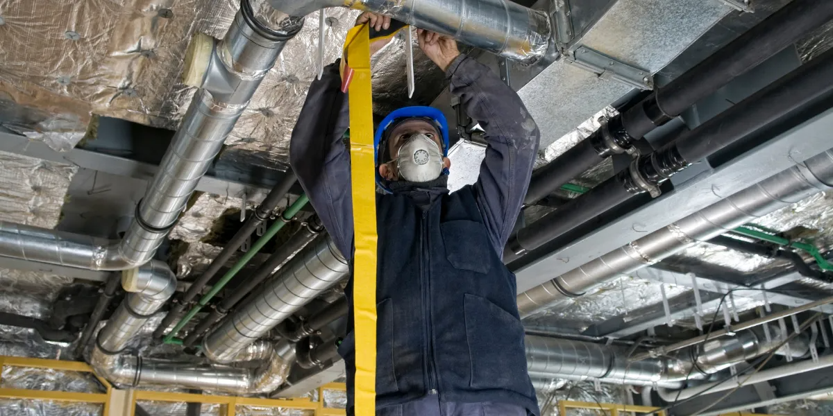 Person in blue hard hat and mask works on pipes overhead.
