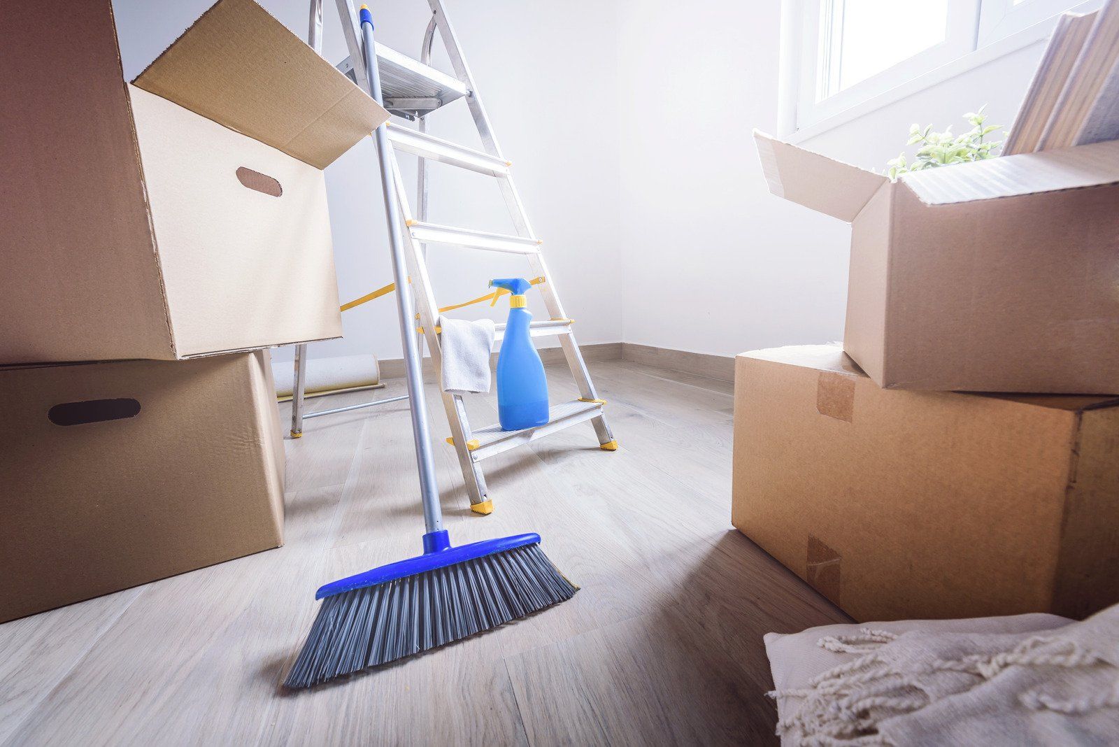Cardboard boxes, a ladder, a broom, and cleaning spray in an empty room, likely preparing for moving.