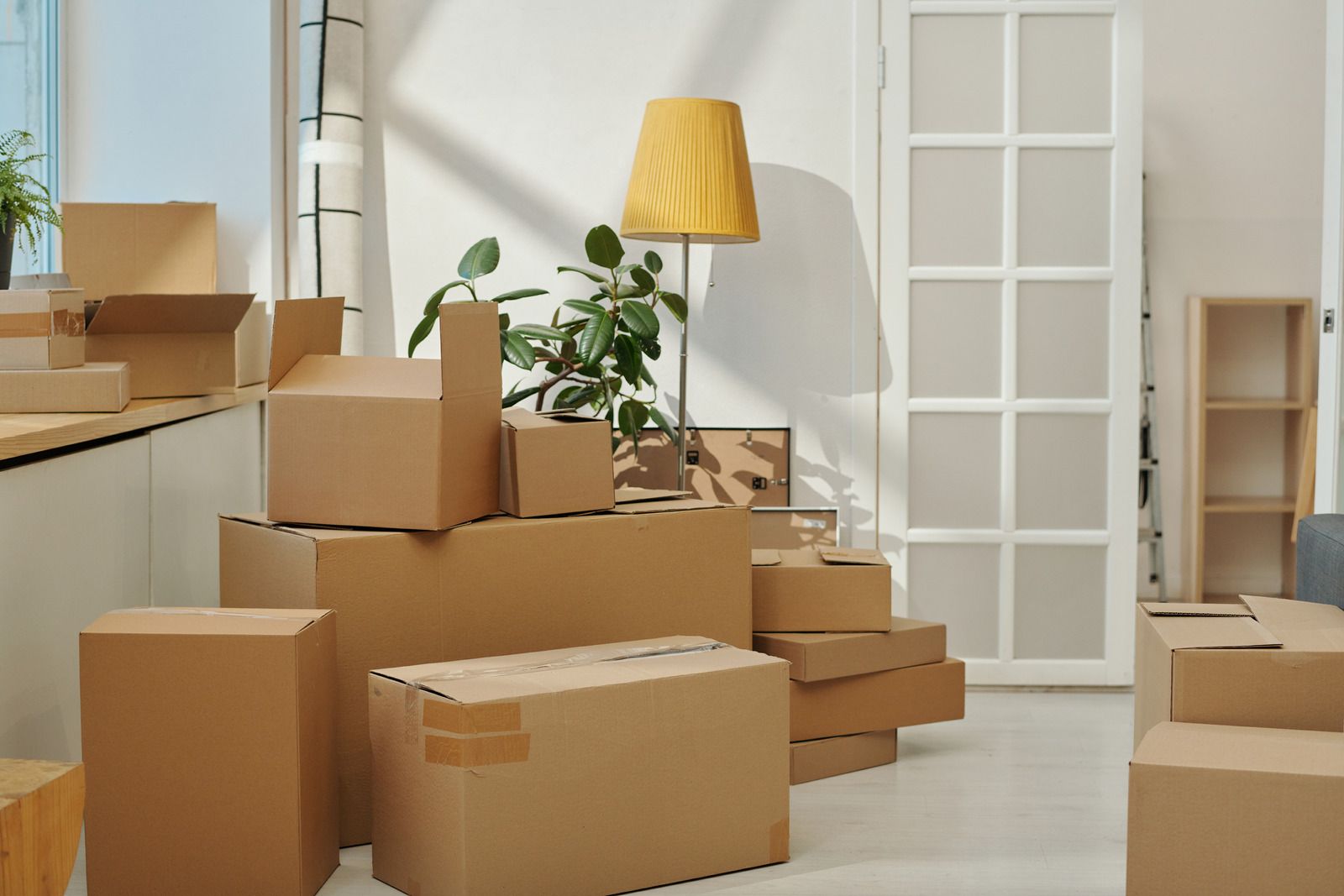 Stacked moving boxes in a bright room with a yellow floor lamp and potted plant