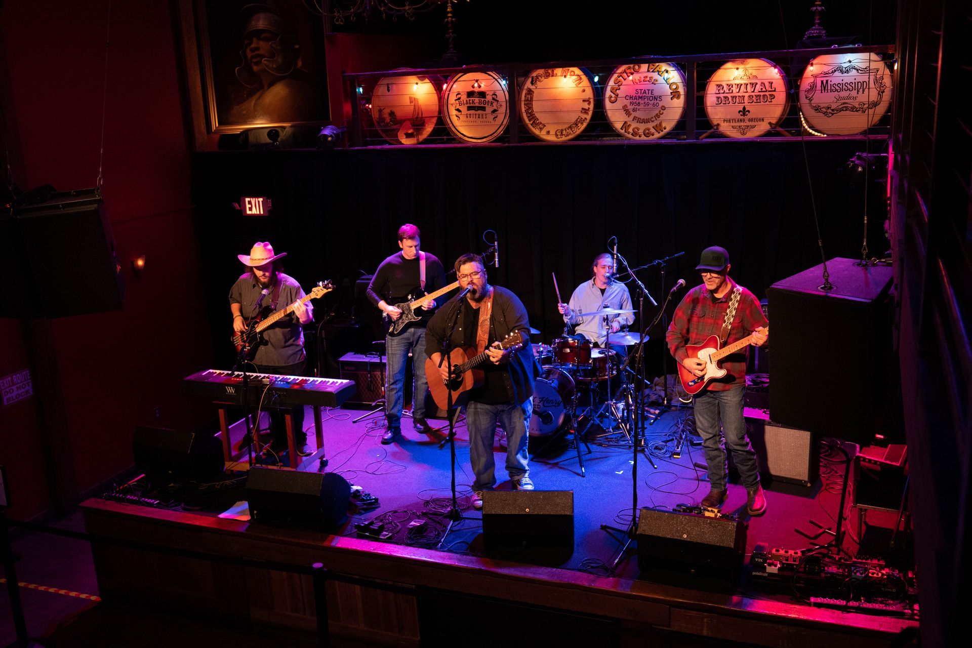 A band of five men performing on a stage with instruments, under stage lights, and with a dark backdrop.