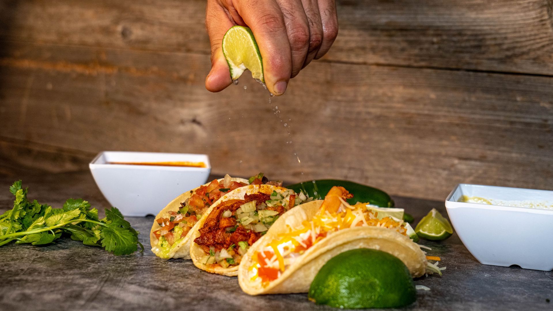 Hand squeezing lime juice over tacos on a table with bowls and lime wedges.