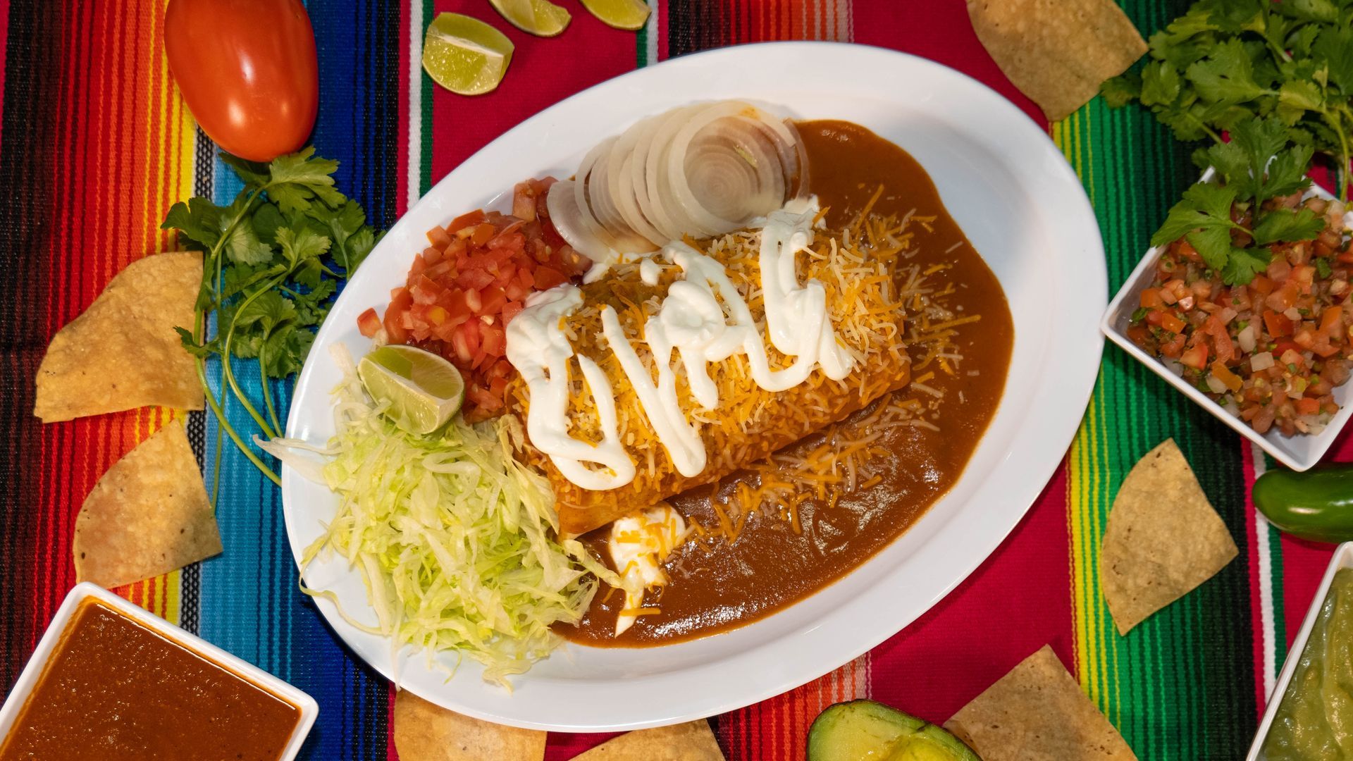 Burrito covered in sauce with toppings on a plate, served with salsa, chips, and garnishes on a colorful tablecloth.