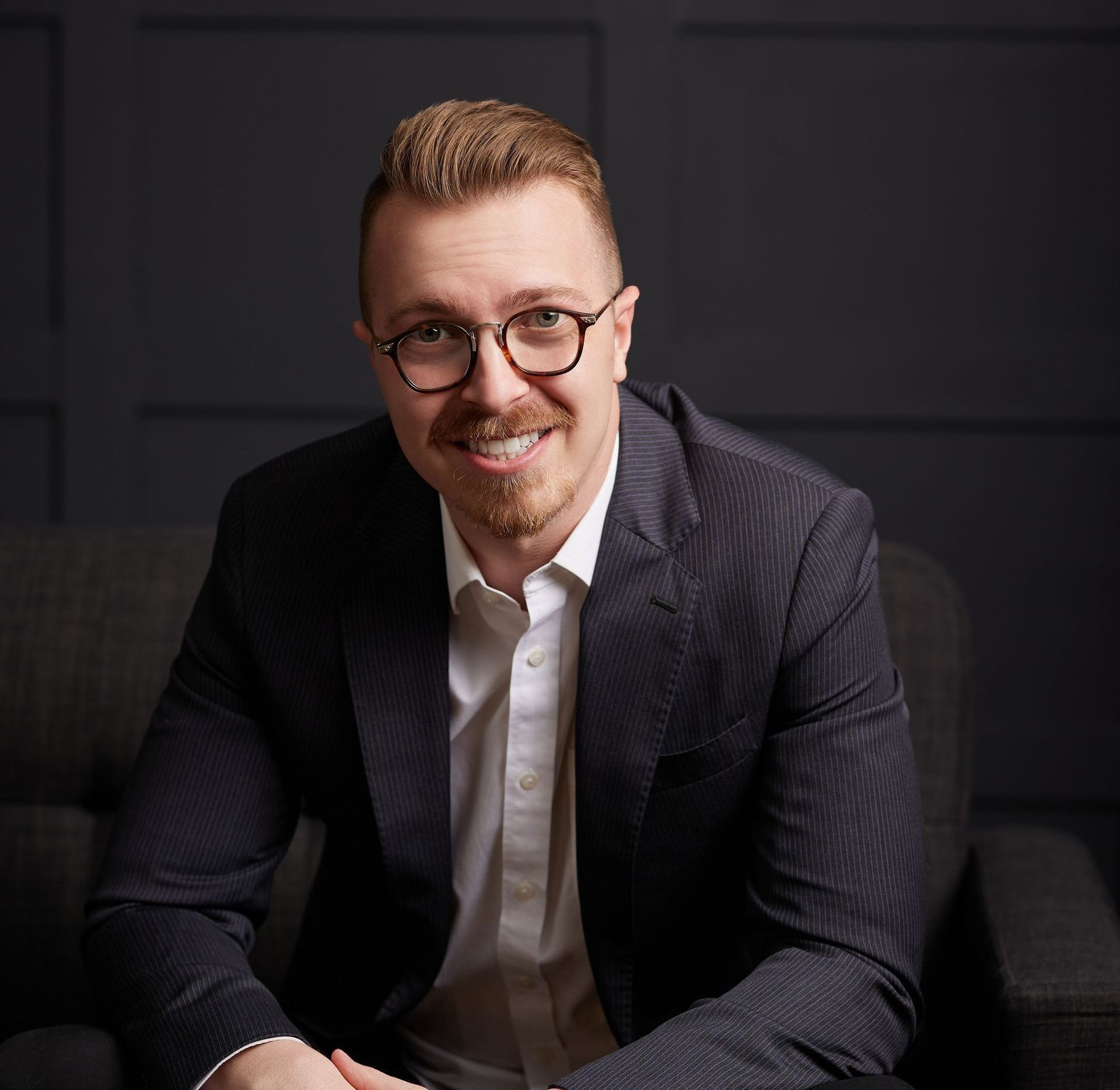 A man in a suit and glasses is sitting in a chair and smiling.