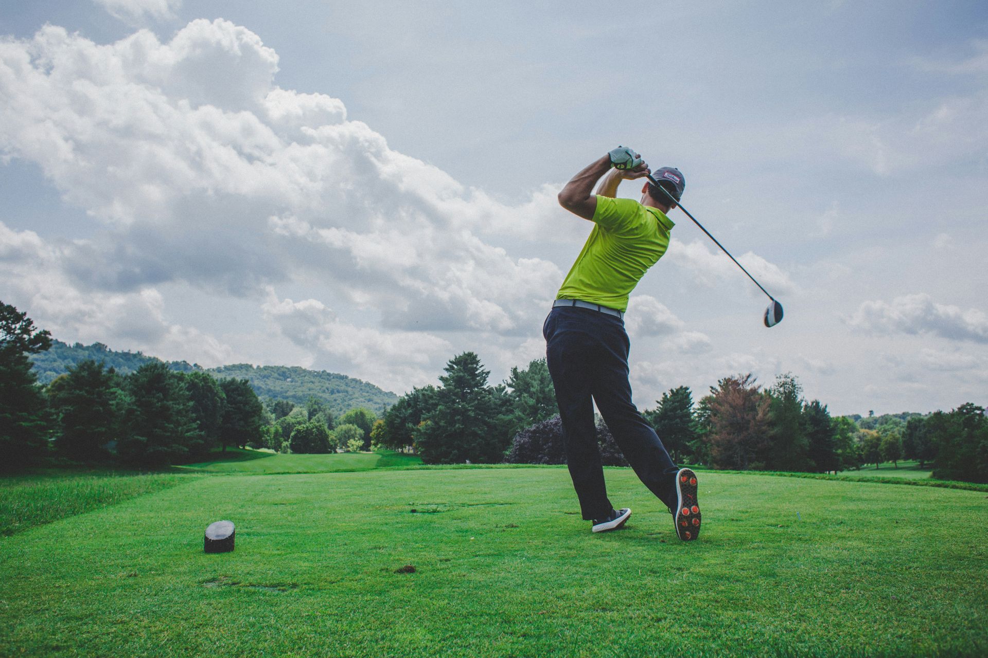 Golfer in neon green shirt swings a club on a grassy tee under cloudy sky.