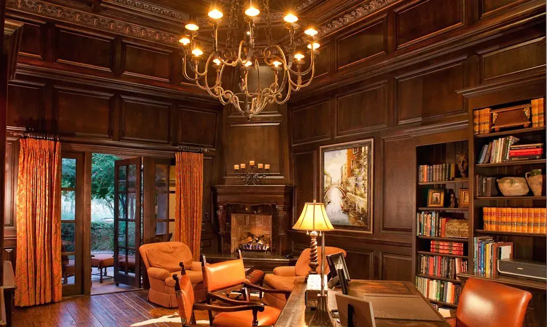 Elegant library with dark wood paneling, fireplace, bookshelves, chandelier, and outdoor view through open doors.