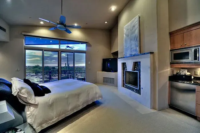 Bedroom with large window overlooking city, bed, fireplace, kitchenette, neutral tones.