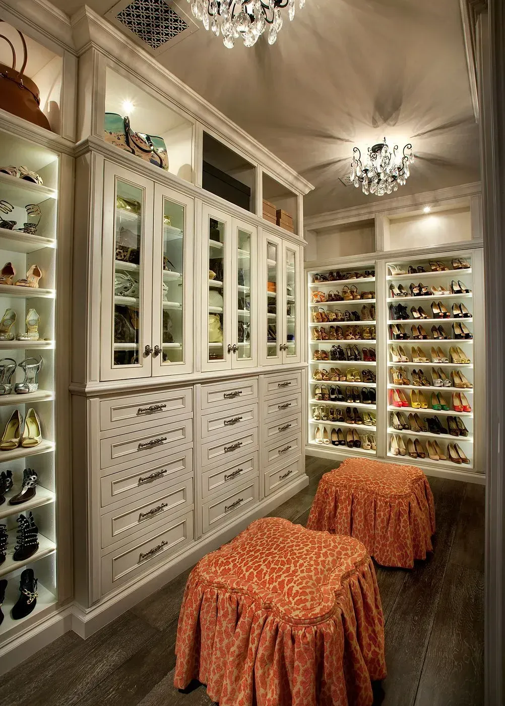 Luxurious walk-in closet with white cabinets, crystal chandeliers, and shoe storage, featuring two orange stools.
