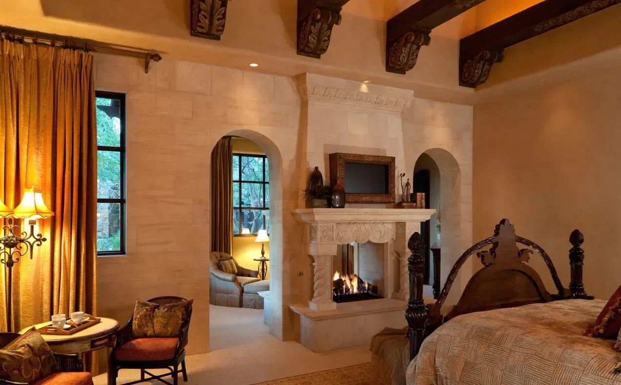 Elegant bedroom with ornate fireplace, arched doorways, and wooden beams. Warm lighting, gold curtains.