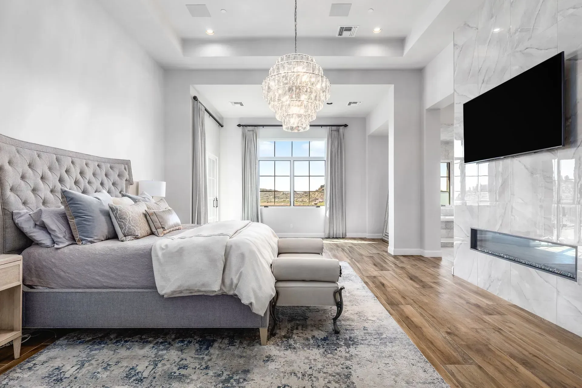 Luxurious bedroom with tufted gray bed, chandelier, fireplace, and large TV.