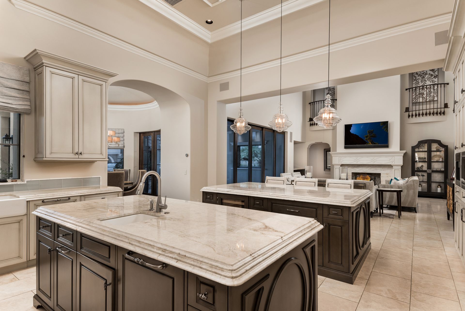 Spacious kitchen with two islands, light countertops, dark cabinets, and pendant lights. A living area is visible in the background.