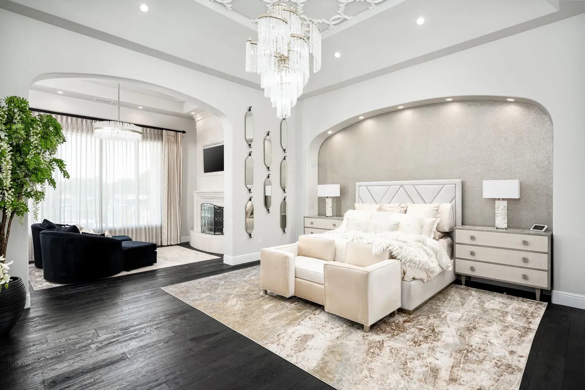 Luxurious bedroom with black wood floors, chandelier, cream furniture, and fireplace.