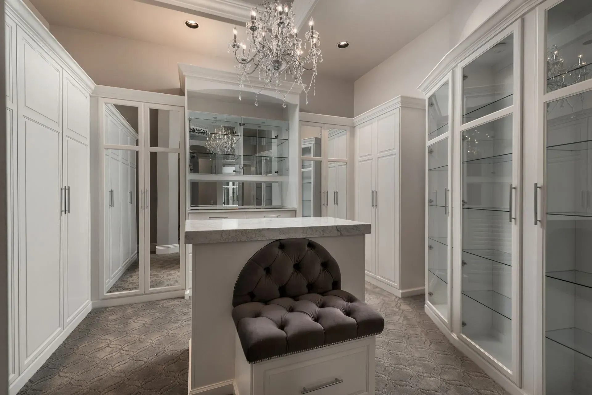 Walk-in closet with white cabinets, glass doors, a tufted seat, and a chandelier.