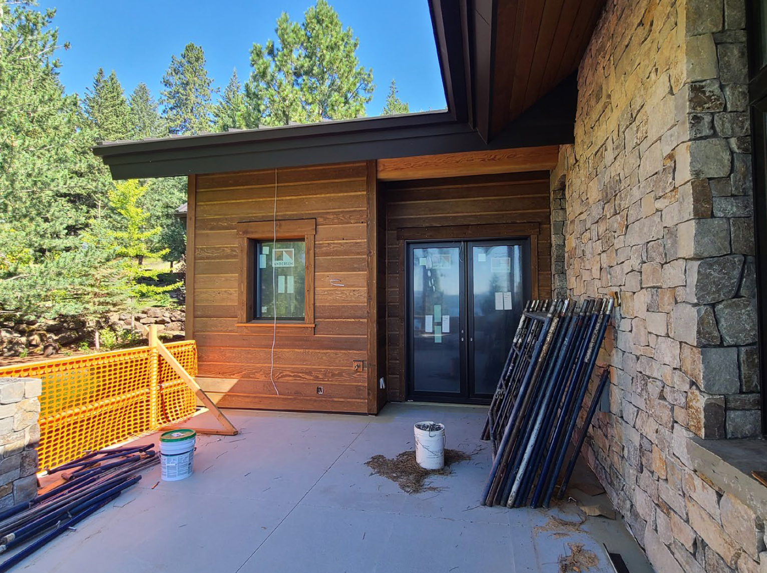 Exterior of a building under construction, featuring wood siding and stone, with glass doors and a yellow safety railing.