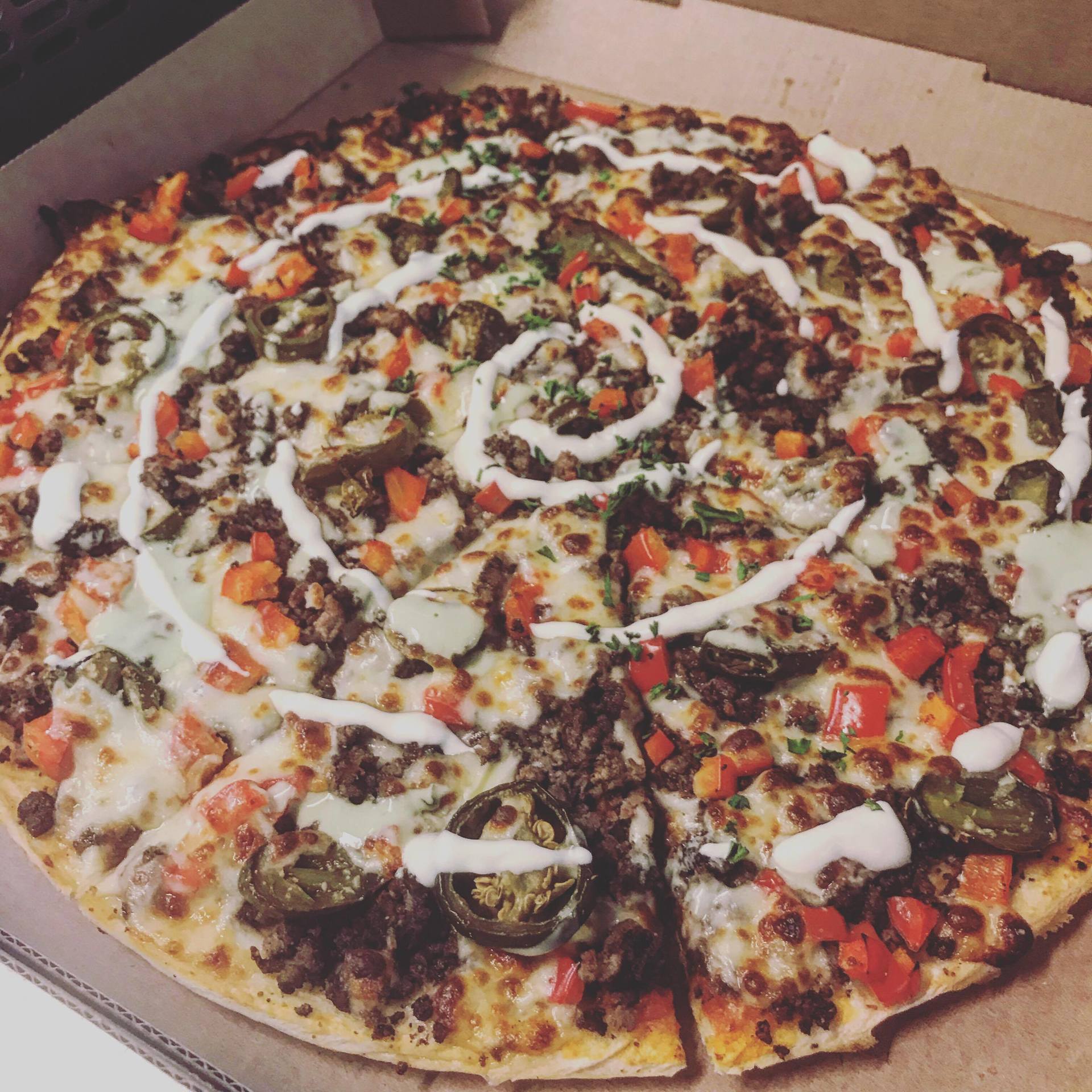 A large pizza in a box, topped with vegetables, meats, and drizzled white sauce.