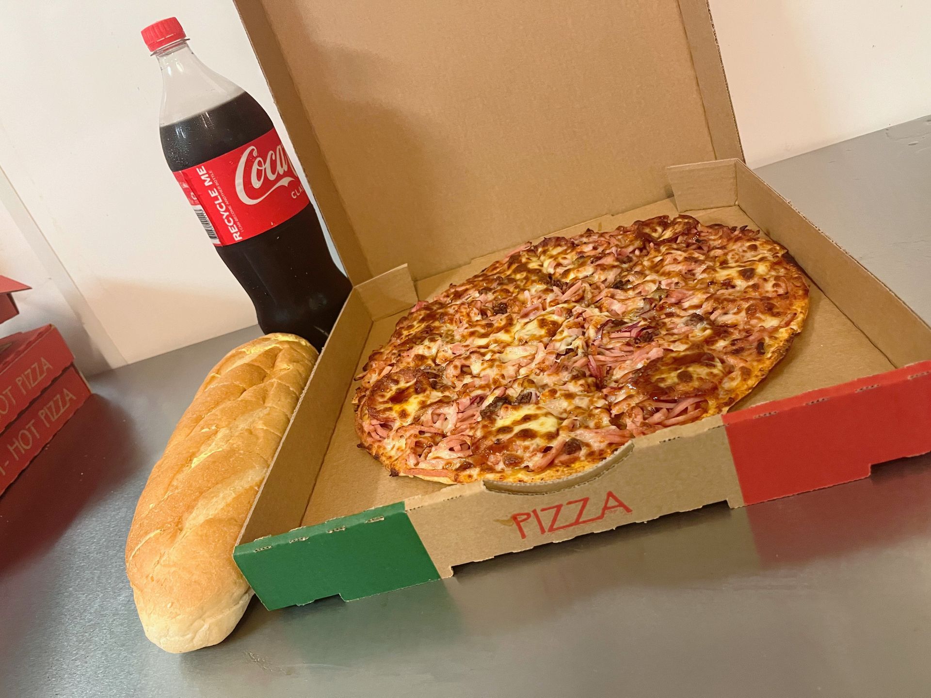 Pizza in an open box with a baguette and a Coca-Cola bottle on a table