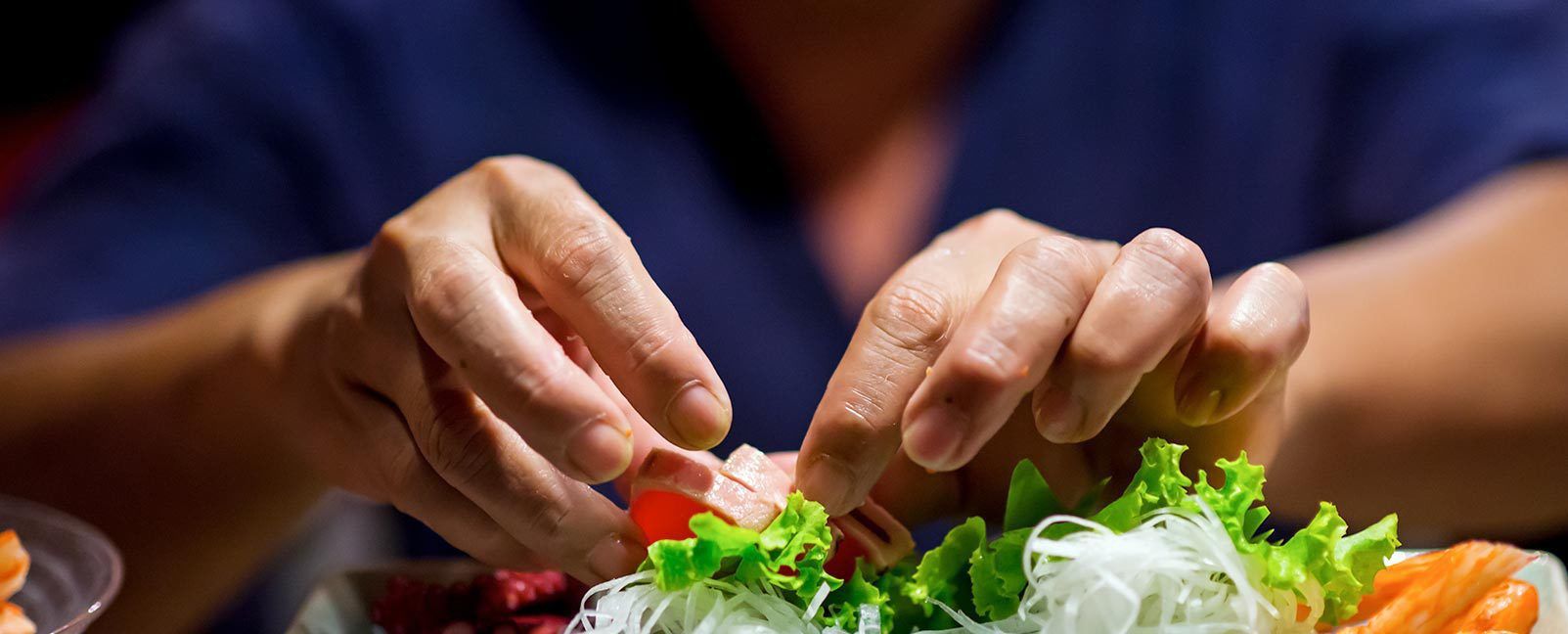 Hands preparing fresh vegetables with a knife on a table, surrounded by leafy greens and tomatoes