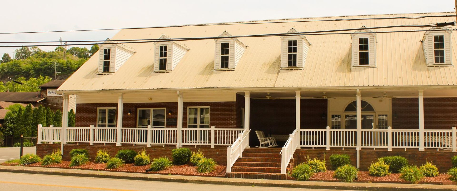 A long, one-story wooden building with a metal roof, dormer windows, and a wide front porch with a white railing.