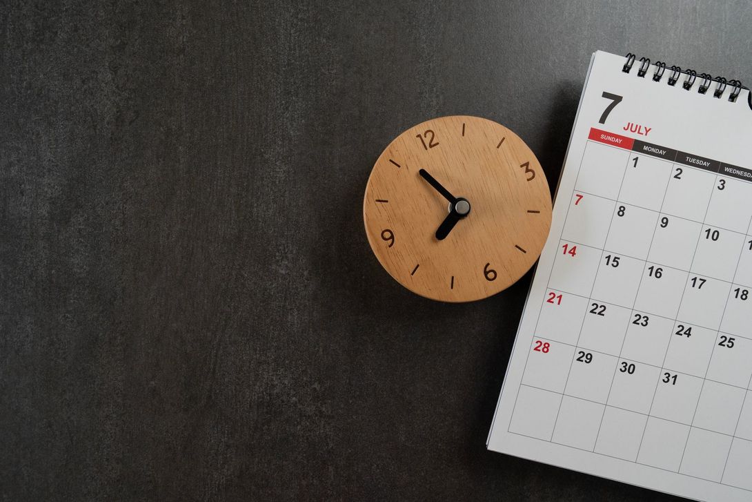A wooden desk clock set to 10:40 sits next to a July calendar on a dark gray surface.