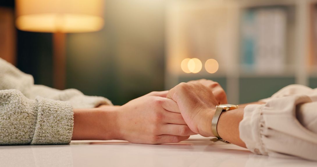 Two people sitting at a table, gently holding hands in a supportive gesture, with a soft-focus warm indoor background.