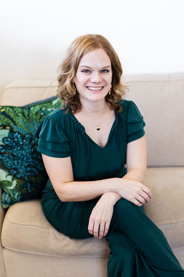 Woman in a dark green outfit sitting on a beige couch, smiling at the camera.