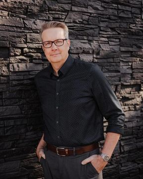 Man standing against a dark stone wall, wearing glasses, a black shirt, and brown pants, hands in pockets.