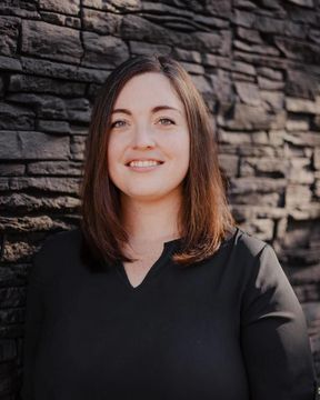 Woman smiling in a black top, standing against a dark stone wall.