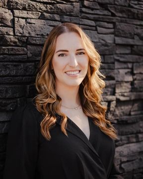 Smiling woman with long wavy hair in a black blouse, standing before a stacked stone wall.