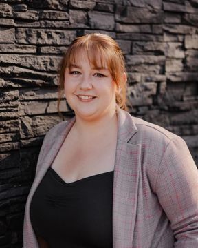 Smiling person in a black top and patterned cardigan standing before a stone wall