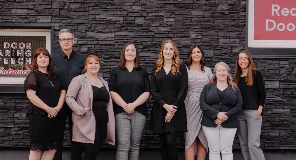 Seven staff members posing outside a red-and-black business storefront, standing in front of a stone wall.