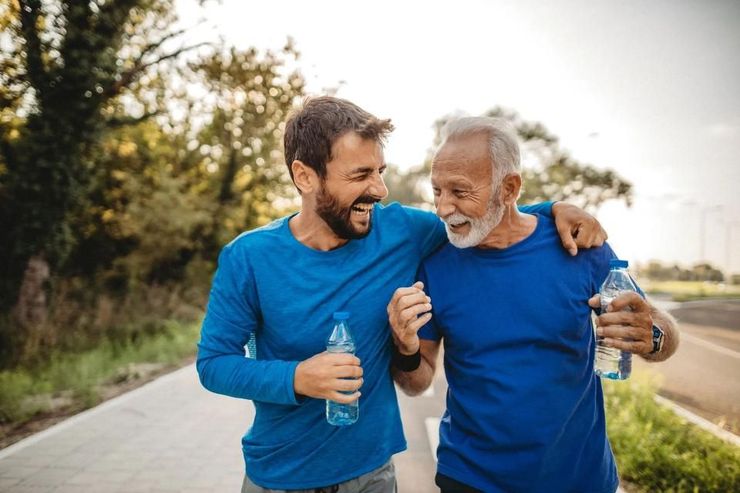 Two men in blue running shirts walk outdoors, smiling and holding water bottles on a tree-lined path.