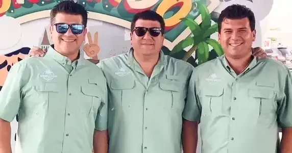 Three men in light green shirts stand together, smiling, with sunglasses in a colorful setting