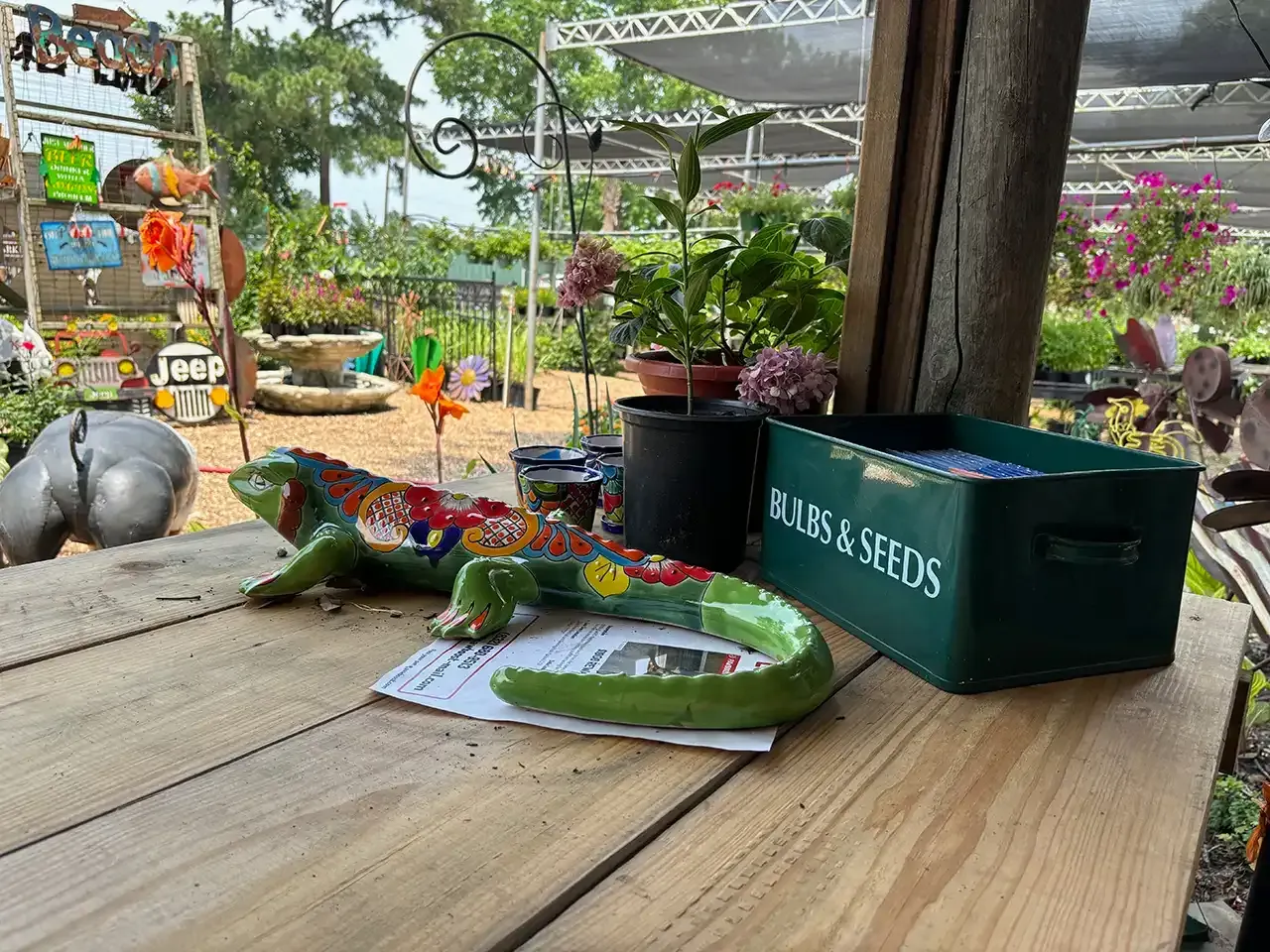 Garden center table with a green seed box, plant pots, and a small lizard figurine on wooden planks.