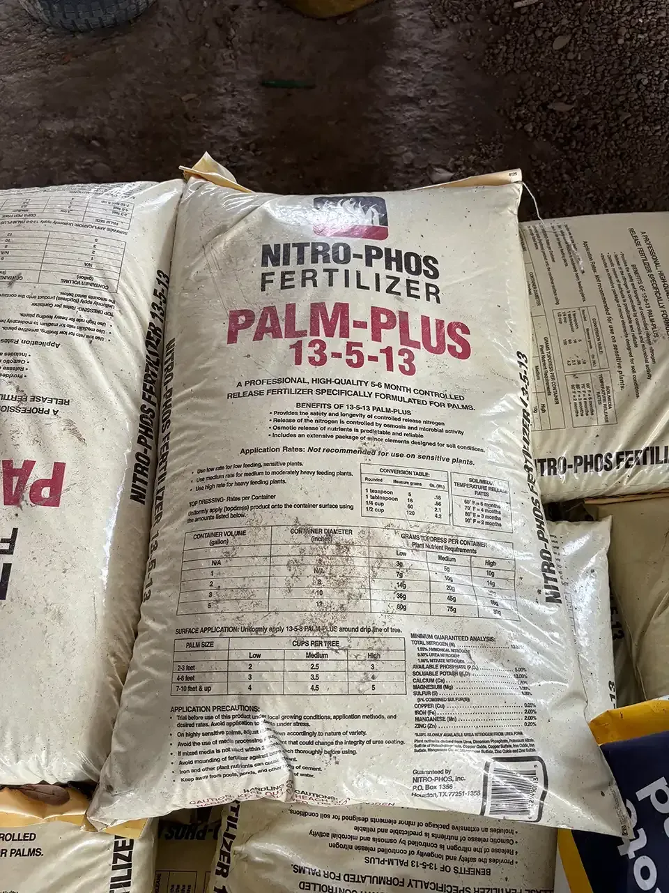 White fertilizer sack labeled “NITRO-PHOS FERTILIZER PALM-PLUS 13-5-13” on a dark floor