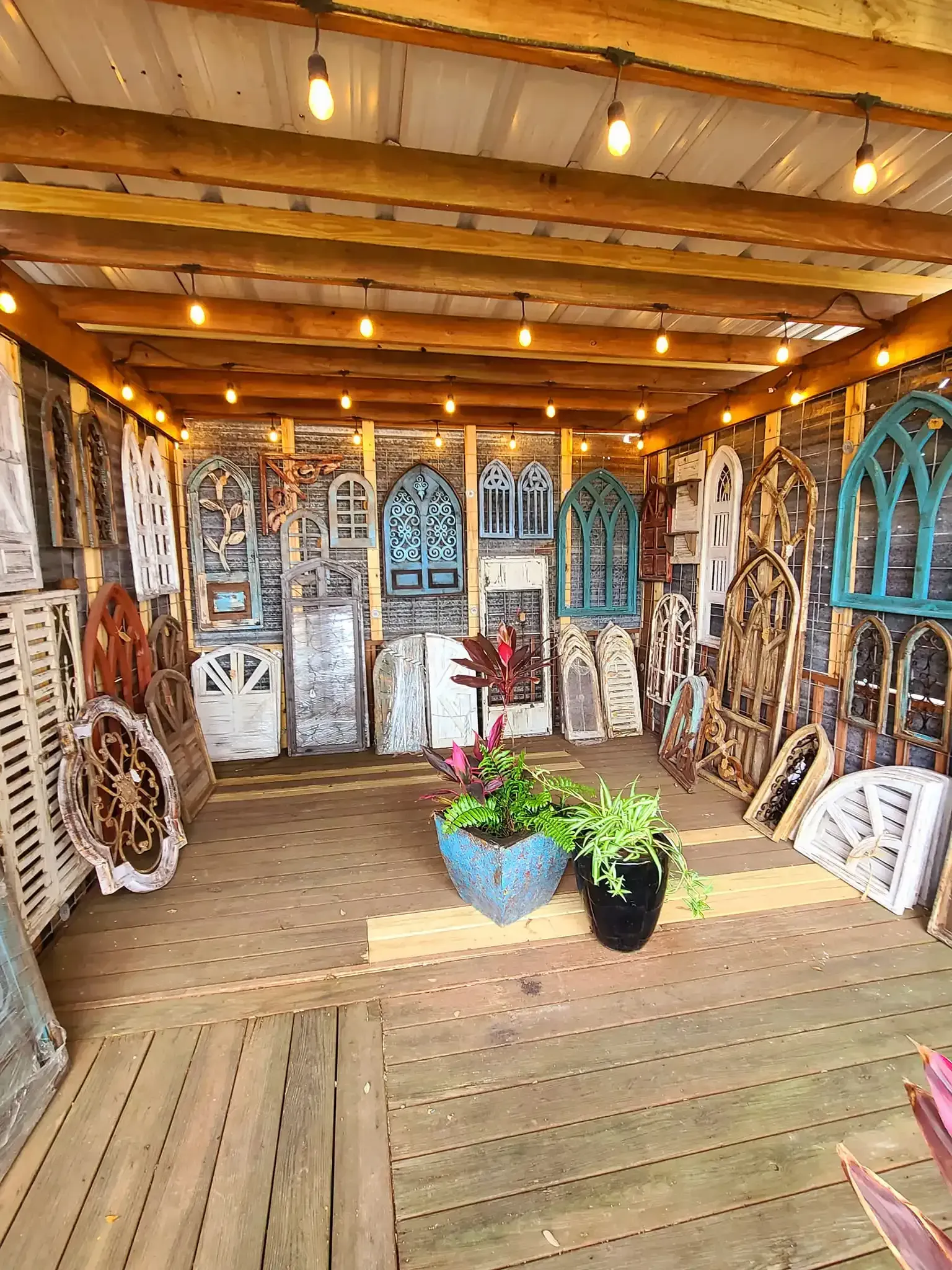 Cozy rustic porch with wood walls, hanging lights, arched windows, and potted plants on a slatted floor