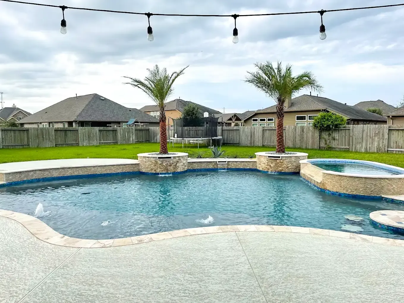 Backyard pool with palm trees, a hot tub, string lights, and a cloudy sky.