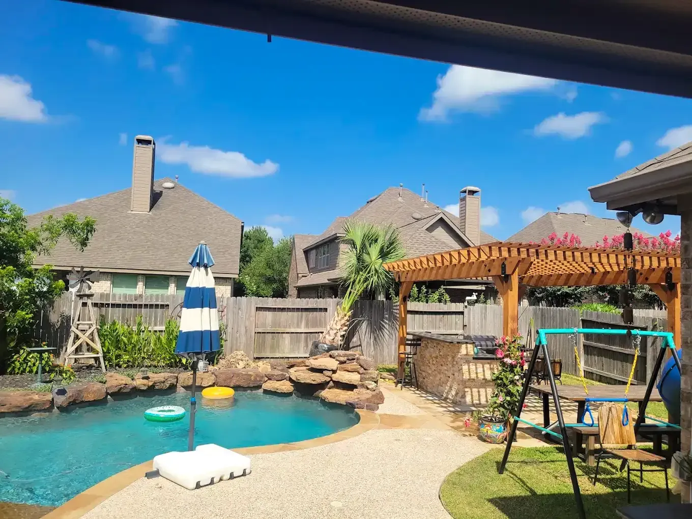 Backyard with pool, pergola, and swing set under a blue sky with fluffy clouds.