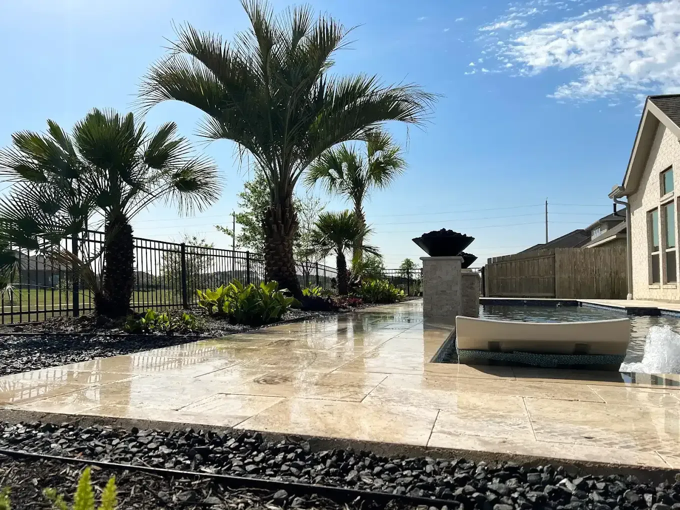Patio with pool, palm trees, and black fence against a blue sky.