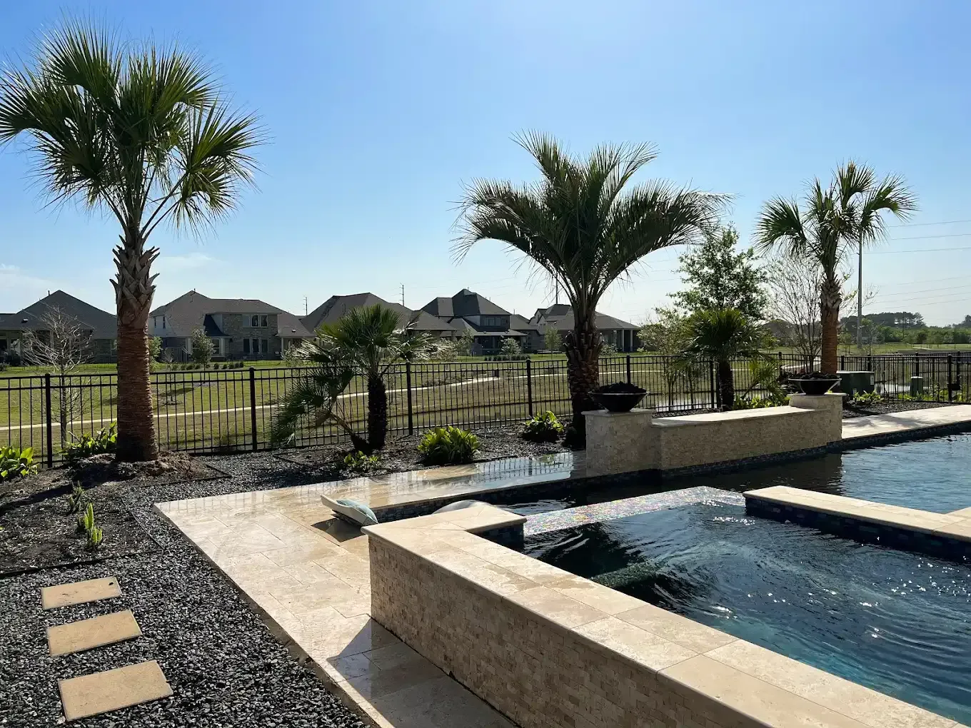 Backyard pool with palm trees, stone patio, and view of neighborhood homes. Blue sky overhead.