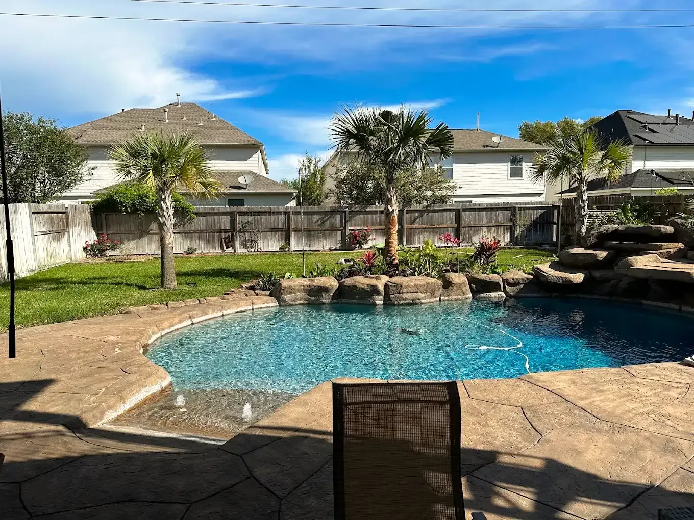 Backyard pool with blue water, patio chairs, and houses beyond a fenced yard.