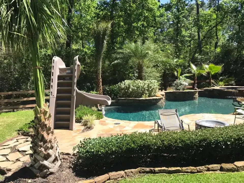 Backyard pool with a small slide, lounge chairs, and lush trees in bright sunlight