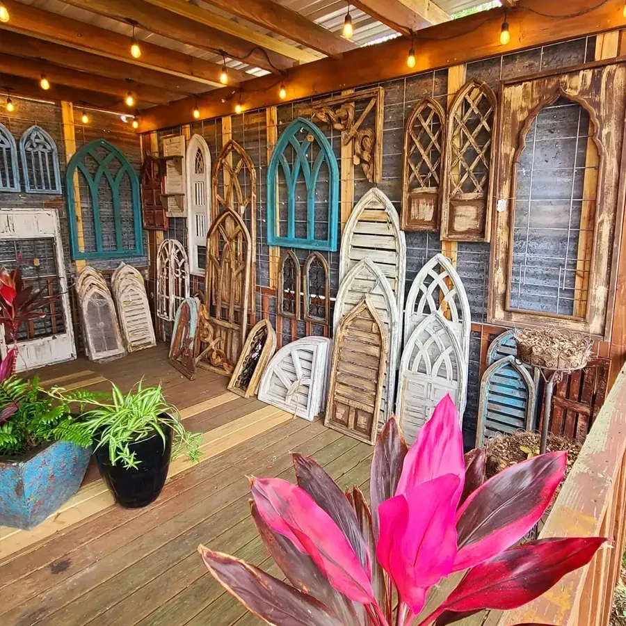 Display of antique arched windows and shutters on a wooden deck, with plants and string lights.