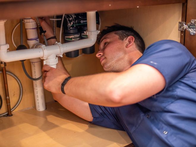 Man kneeling under a kitchen sink, repairing plumbing with a flashlight and tools on the floor
